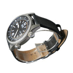 A wristwatch with a black dial, silver-toned metal case, and black leather strap, displayed on a clear stand against a gray background.