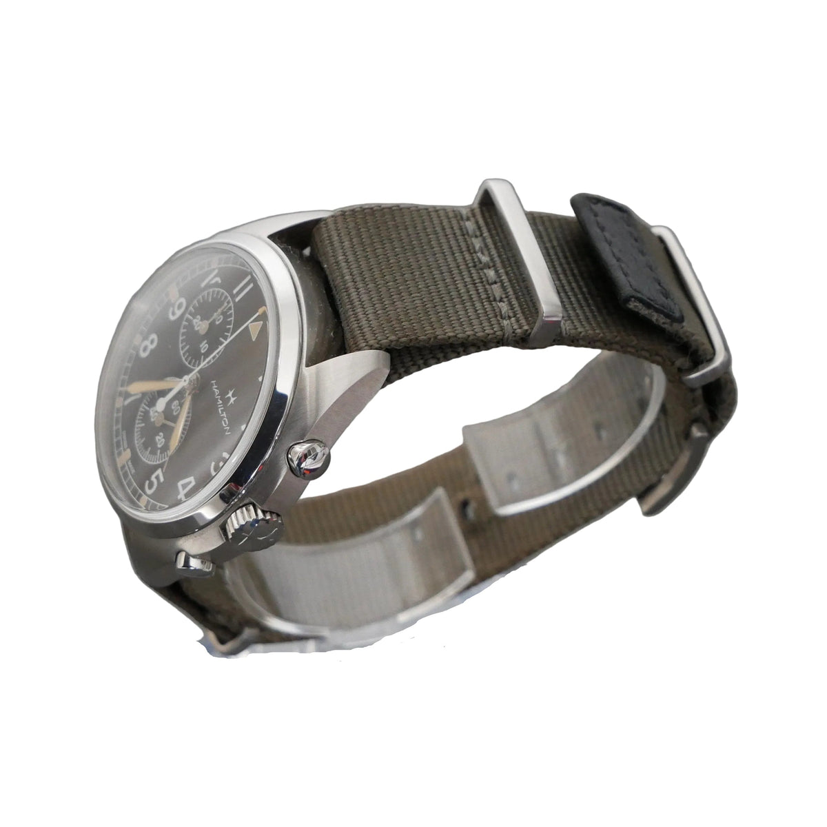 A wristwatch with a silver-toned metal case, a dark gray dial, and an olive green canvas strap, displayed on a clear acrylic stand against a dark gray textured background.