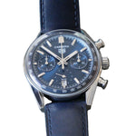 A Carrera chronograph watch with a dark blue dial and a dark blue leather strap, resting on a textured gray surface. The watch features a silver-tone case with a tachymeter bezel and a silver-tone day-date window.