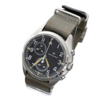 Hamilton watch with a black dial, silver-tone stainless steel case, and an olive green canvas strap, displayed on a clear acrylic stand against a gray background.