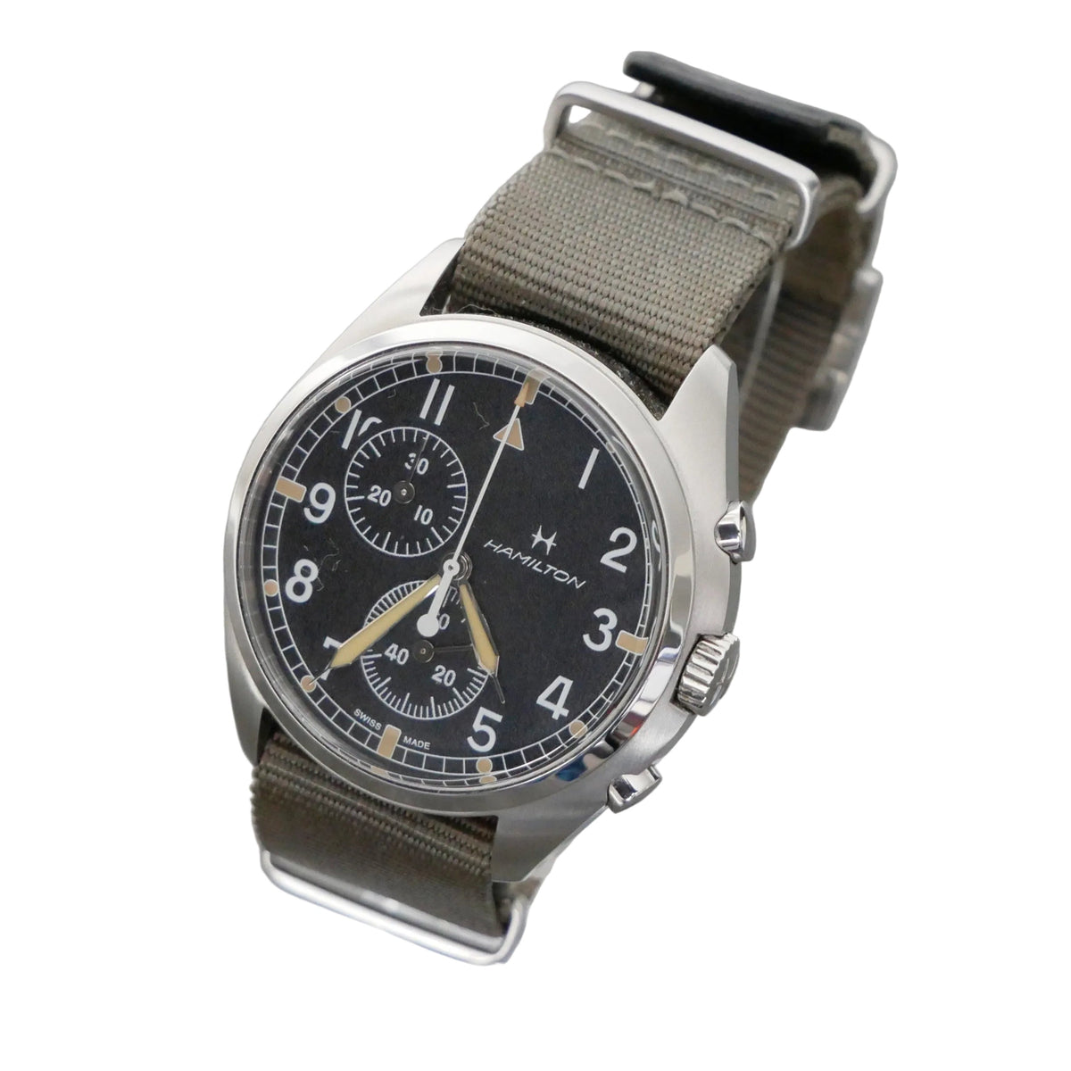 Hamilton watch with a black dial, silver-tone stainless steel case, and an olive green canvas strap, displayed on a clear acrylic stand against a gray background.