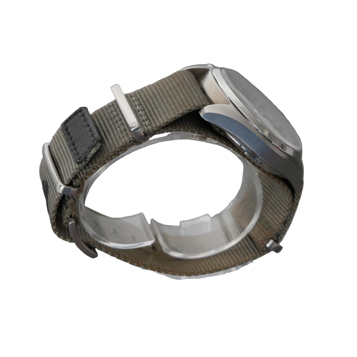 A silver-tone analog watch with an olive green woven nylon strap, displayed on a clear acrylic stand against a textured gray background.