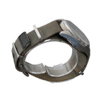 A silver-tone analog watch with an olive green woven nylon strap, displayed on a clear acrylic stand against a textured gray background.